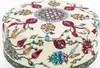 A close-up view of a round ottoman with a cream-colored fabric featuring a colorful floral and geometric pattern. Produktbild-Vorschau