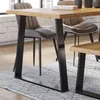 A close-up view of a dining table with black trapezoidal legs, a wooden tabletop, and upholstered chairs in a dining room setting. Náhľad obrázku produktu