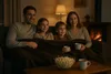 A family is watching TV by the fireplace, covered by a blanket, with popcorn and mugs. Produktbild-Vorschau