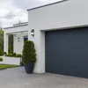 A modern house with a dark gray garage door and white exterior walls, featuring a tall potted evergreen tree in front of the garage. Anteprima dell’immagine prodotto
