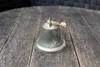 A small metal bell with a rope handle sits on a wooden surface. Anteprima dell’immagine prodotto