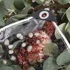 A close-up view of a transparent tarp with a grommet and rope, partially covering a floral arrangement with eucalyptus leaves. Náhled obrázku produktu