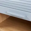 A close-up view of a light gray cabinet with a roller shutter door and a wooden shelf below. Produktbild-Vorschau
