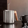 A stainless steel teapot with a wooden handle sits on a dark wooden table next to a black mug. Náhled obrázku produktu