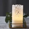 A white LED candle with a decorative filigree design sits on a mirrored tray with a brass frame, next to some green foliage. Podgląd zdjęcia produktu