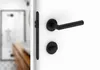 A close-up shot of a black door handle and lock set on a white door, with a blurred view of a bathroom in the background. Produktbild-Vorschau