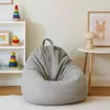 A light gray bean bag chair sits on a white rug in a child's room, next to a white shelf with toys and a small round table with a mushroom lamp and a framed picture. Náhled obrázku produktu