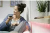 A woman is sitting on a couch and holding a smartphone, looking away from the camera. Podgląd zdjęcia produktu