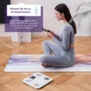A woman is sitting on a yoga mat, looking at her phone, with a smart scale in the foreground. Anteprima dell’immagine prodotto