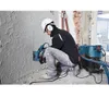 A construction worker is using a Bosch angle grinder with a dust extraction hood attached to cut into a textured wall, with a vacuum cleaner connected. Voir les images du produit