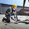 A construction worker is kneeling and using an angle grinder to cut a metal beam on a concrete surface. There are stacks of metal rods in the background. Náhľad obrázku produktu