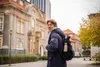A young man wearing a blue winter coat and a backpack is standing on a street in front of a building. Náhled obrázku produktu