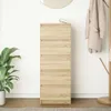 A tall, narrow chest of drawers with a light wood grain finish is shown against a white wall. A large green plant in a white pot is to the left, and a coat rack with a beige coat is to the right. A round mirror is visible at the top. Náhled obrázku produktu
