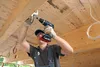 A person is using a cordless drill to screw something into a wooden beam on a ceiling. Voir les images du produit