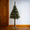 A decorated artificial Christmas tree with a real wood trunk and jute sack base, standing indoors next to a wooden door. Produktbild-Vorschau