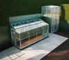 Two greenhouses are shown in an outdoor setting, one larger and one smaller and taller, against a green hedge and fence. Náhled obrázku produktu