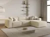A modern beige sectional sofa is shown in a well-lit living room setting, with shelves on the wall behind it and a coffee table in front. Produktbild-Vorschau