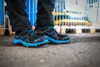 A person wearing black pants and black and blue safety shoes stands on a concrete surface, with a blurred industrial background. Náhľad obrázku produktu