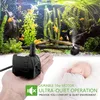 A hand holding a small black water pump in front of a fish tank, with a sleeping baby in the background. Produktbild-Vorschau