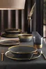 A table setting with dark gray and gold-rimmed dinnerware, a glass, and a vase with dried grass. Produktbild-Vorschau