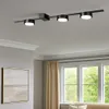 A track light fixture mounted on a ceiling in a room with a painting on the wall and windows with curtains. Produktbild-Vorschau