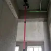 A red telescopic pole with a laser level attached to the ceiling, projecting a green laser line. Náhled obrázku produktu