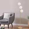 A modern floor lamp with three white globe shades stands next to a grey armchair with a white textured pillow. Books are stacked on the floor in front of the chair. Podgląd zdjęcia produktu