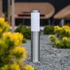 A silver bollard light with a white diffuser stands on a gravel path, surrounded by yellow and green foliage. Anteprima dell’immagine prodotto