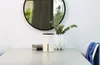 A white cylindrical object with a black top sits on a gray table next to a vase with green leaves and a stack of books. A round mirror hangs on the white wall behind the table. Náhled obrázku produktu
