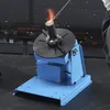 A welding positioner is shown in use, with sparks flying from the welding rod. A person's legs are visible on the right side of the image. Podgląd zdjęcia produktu