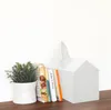 A white house-shaped tissue box sits on a wooden surface next to a small potted plant and a stack of books. Anteprima dell’immagine prodotto