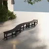 A long, curved wooden bench sits on a tiled patio under a tree. Náhled obrázku produktu