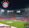A football stadium filled with fans holding up red and white cards to create a mosaic of the FC Bayern München logo, with the team's logo and stars in the upper left corner. Produktbild-Vorschau