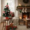 A decorated Christmas tree sits on a side table next to a fireplace with stockings hanging. Náhled obrázku produktu