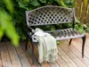 A brown metal garden bench with a green throw blanket and a book on it, placed on a wooden deck with lush green foliage in the background. Náhľad obrázku produktu
