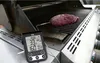 A digital thermometer is shown inserted into a raw steak on a gas grill. The thermometer displays temperature readings and other data. Produktbild-Vorschau