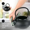 A person is drying a black cast iron teapot with a green cloth, with an inset showing the teapot being rinsed under a faucet. Produktbild-Vorschau