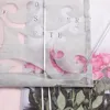 A close-up view of a gray fabric blind with a floral pattern and text. The blind is partially unrolled, revealing pink roses and green leaves underneath. A clear plastic rod is visible along the right edge. Produktbild-Vorschau