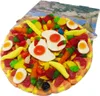 A close-up of a gummy candy pizza with various fruit-flavored gummy toppings, including gummy eggs, carrots, strawberries, and bananas. The pizza is on a white background, with a blurred image of a coastal town in the background. Produktbild-Vorschau