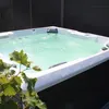 An outdoor whirlpool filled with bubbling green water, with a dark fence in the background and some green leaves in the foreground. Podgląd zdjęcia produktu