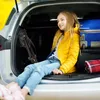A young girl sits in the trunk of a car with luggage. Produktbild-Vorschau