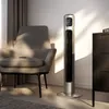 A modern silver tower fan is placed in a living room next to an armchair and a dresser. Sunlight streams in from the side, creating dramatic shadows. The fan has a digital display showing '22'. Náhled obrázku produktu