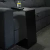 A black C-shaped side table is placed next to a dark gray sofa, with a glass of water and a remote control on top of it. Produktbild-Vorschau