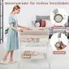 A woman is standing next to a baby changing table with a baby on it. The table has storage underneath and wheels. There are inset images showing the wheels. Produktbild-Vorschau