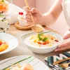 A person is eating fruit salad from a bowl with a gold spoon, with other plates of food and drinks on a table. Náhled obrázku produktu