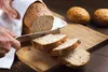 A person is slicing a loaf of bread on a wooden cutting board, with two small buns in the background. Náhľad obrázku produktu