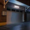 A linear LED light fixture illuminates a brick wall with two garage doors at night. Produktbild-Vorschau