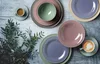 A collection of ceramic plates and bowls in various pastel colors, arranged on a rustic wooden surface with some greenery. Produktbild-Vorschau