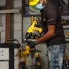 A person wearing a hard hat and face mask is using a yellow and black Dewalt rotary hammer drill to break a brick wall. Other Dewalt tools are visible in the background. Produktbild-Vorschau