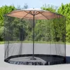 A mosquito net is draped around a patio umbrella, which is set up outdoors on a paved surface with trees and foliage in the background. Produktbild-Vorschau